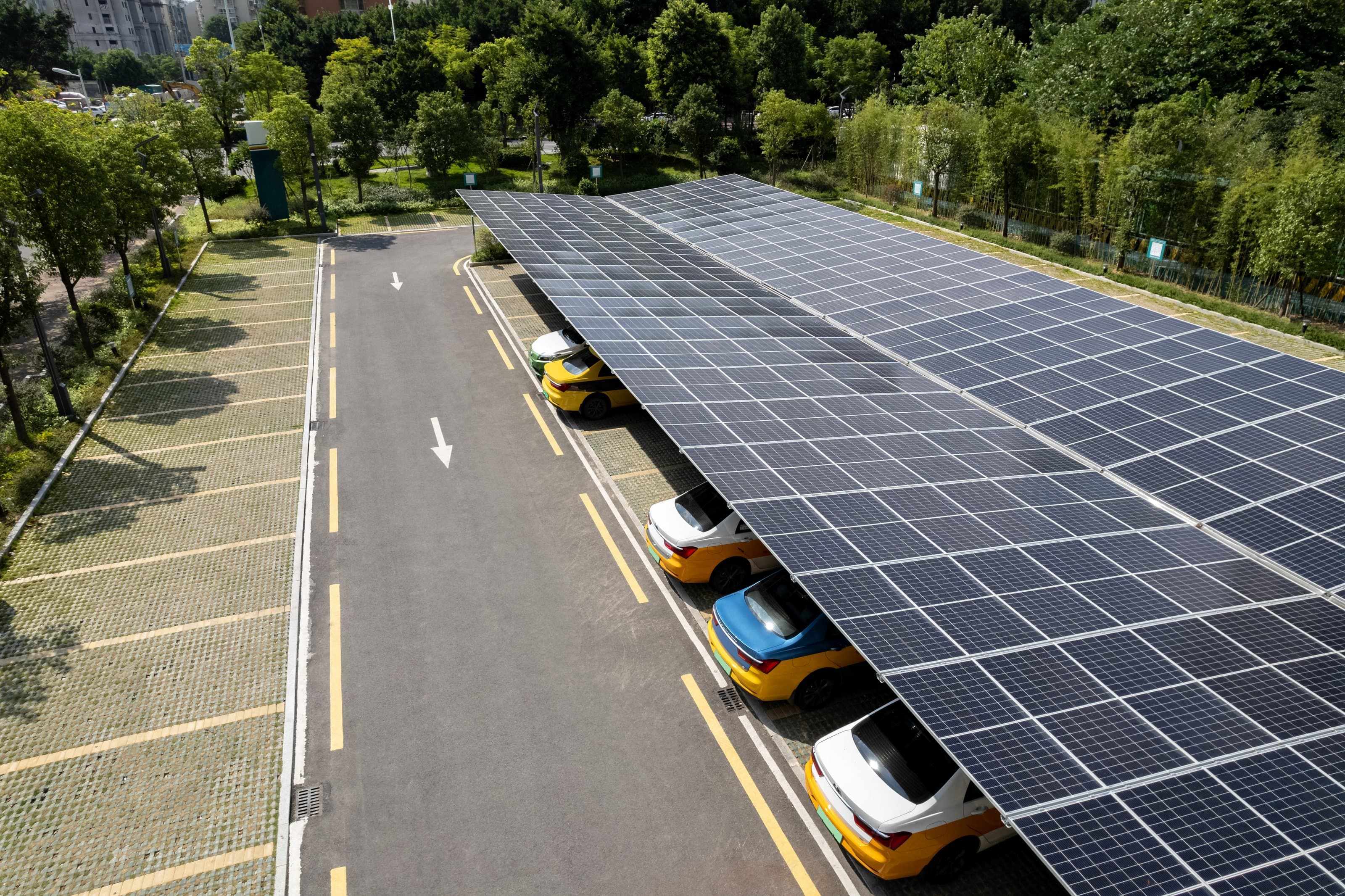 Drone view of a single-row solar carport shading a line of parked cars beside a landscaped lot